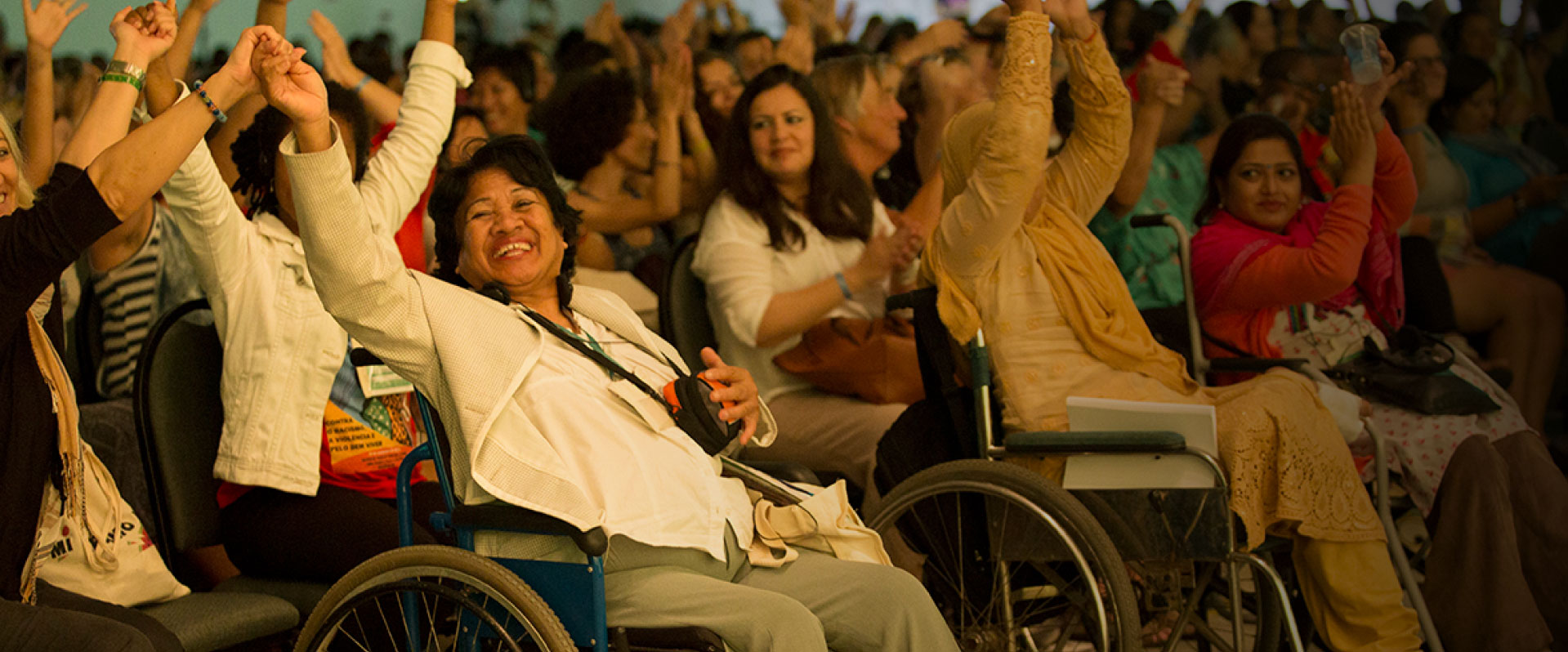 La photo montre un auditorium avec une première rangée de personnes assises dans des fauteuils roulants ayant toutes les mains levées, quelques-unes sont sur des chaises dans le coin de la photo et d’autres sont en train d’applaudir. Hybrid Forum