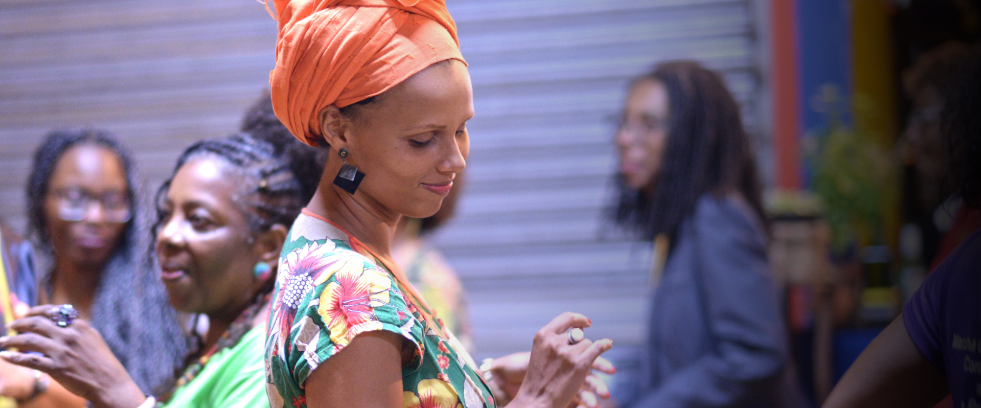 The image centers a person wearing an orange turban, black earrings and a green half-sleeve shirt with yellow and red flowers. They appear to be dancing while smiling as they look towards their right shoulder. The photo also includes three other individuals slightly blurred in the background who also appear to be dancing and/or standing. Register Now