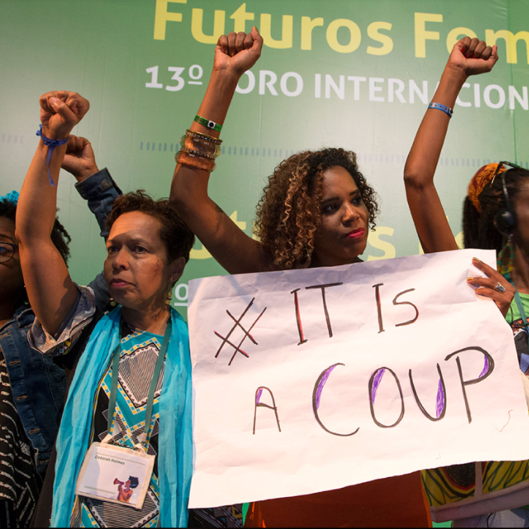The image shows a group of people holding a sign with the text “ # IT IS A COUP." The individuals appear to be serious and standing their ground, and it appears to be a demonstration or protest. The group appear to be standing in front of a banner from the 13th AWID International Forum, written in Spanish. The image shows a group of people holding a sign with the text “ # IT IS A COUP." The individuals appear to be serious and standing their ground, and it appears to be a demonstration or protest. The group appear to be standing in front of a banner from the 13th AWID International Forum, written in Spanish.