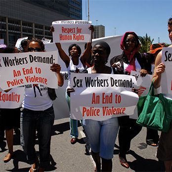The image shows a group of people holding signs in black and red during a protest by, with and for sex workers. The signs display demands related to respecting sex workers’ rights and demands to end police violence, and supporting their human rights. The protestors are standing outdoors under a clear blue sky, wearing casual clothing such as t-shirts, jeans and cargo pants and smiling confidently as they hold up their demands. The image shows a group of people holding signs in black and red during a protest by, with and for sex workers. The signs display demands related to respecting sex workers’ rights and demands to end police violence, and supporting their human rights. The protestors are standing outdoors under a clear blue sky, wearing casual clothing such as t-shirts, jeans and cargo pants and smiling confidently as they hold up their demands.